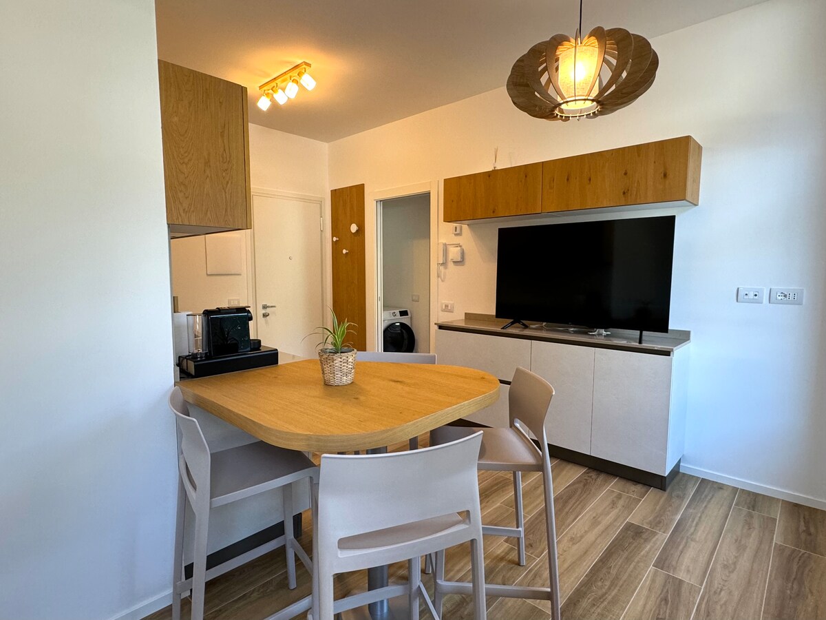 A modern dining area is highlighted by a round wooden table surrounded by four light-colored chairs. A wall-mounted television is positioned across from the table, while overhead lighting provides illumination. The space features neutral-toned cabinetry and a sleek coffee maker on the countertop.