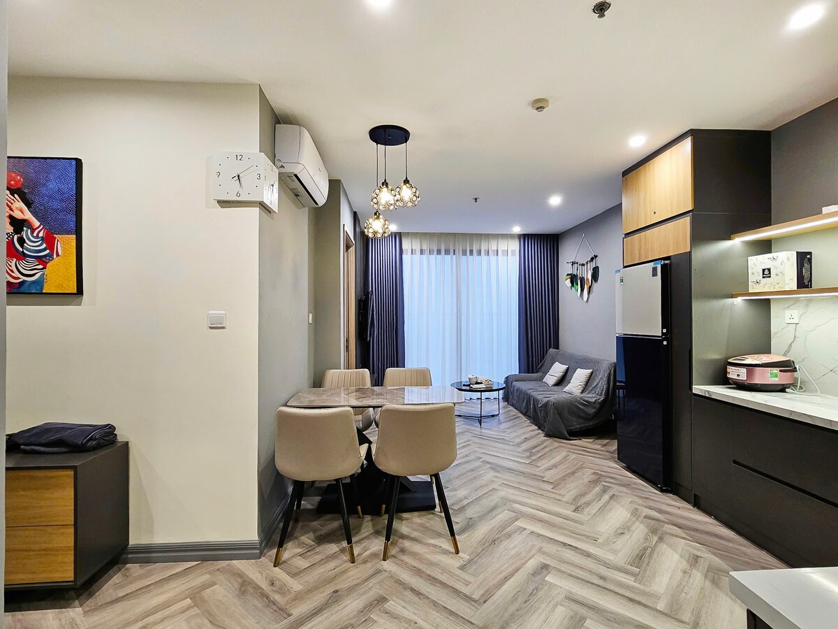 A modern living space is visible, featuring a dining table with four chairs and a cozy seating area with a sofa. Natural light filters through sheer curtains, illuminating the wood-patterned floor. Decorative elements and a sleek kitchen area are included, enhancing the overall appeal.