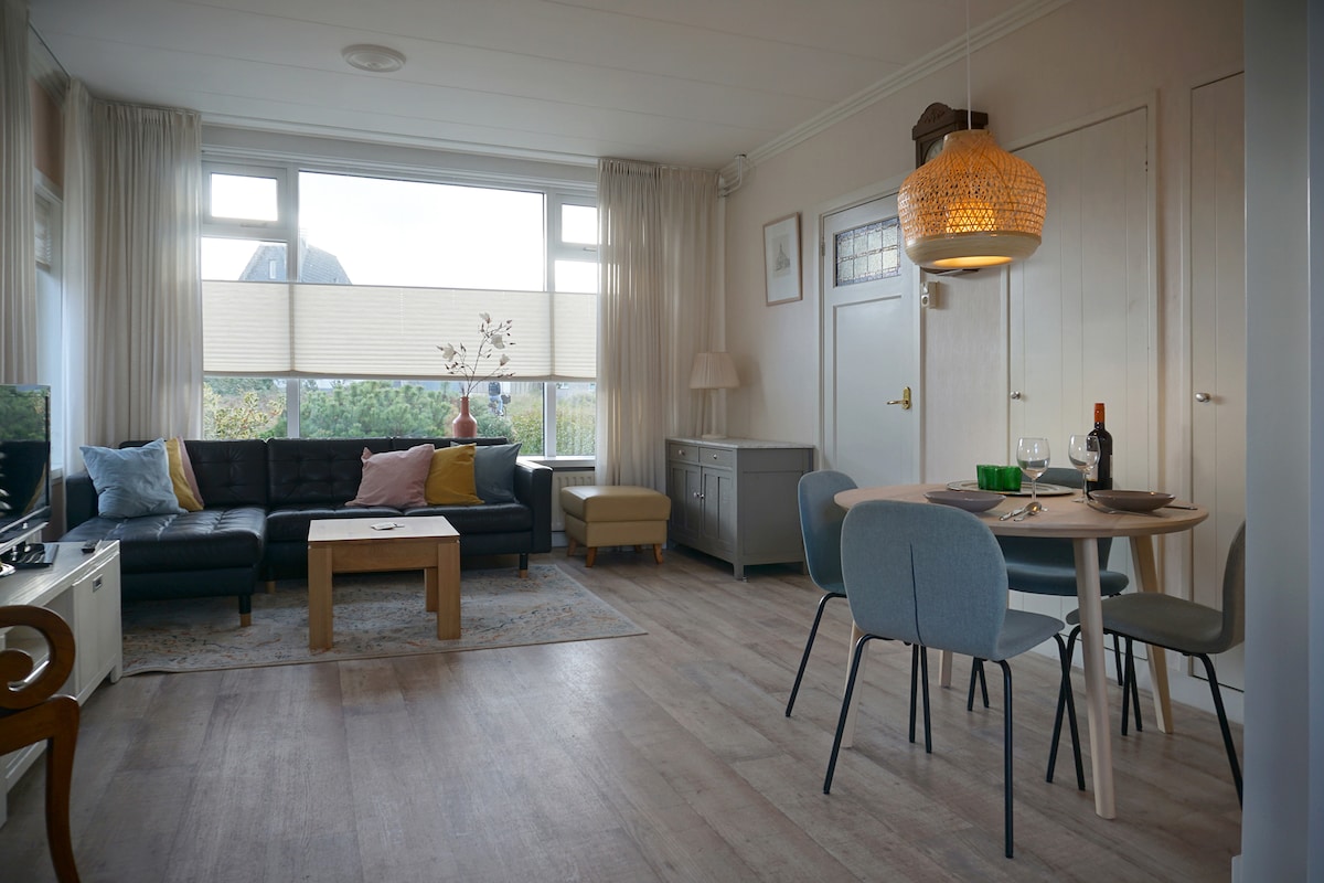A welcoming living area features a comfortable dark sofa adorned with colorful cushions opposite a light wooden coffee table. A dining table with chairs is situated nearby, illuminated by a woven pendant light. Expansive windows offer a view of the outdoors, enhancing the airy ambiance.