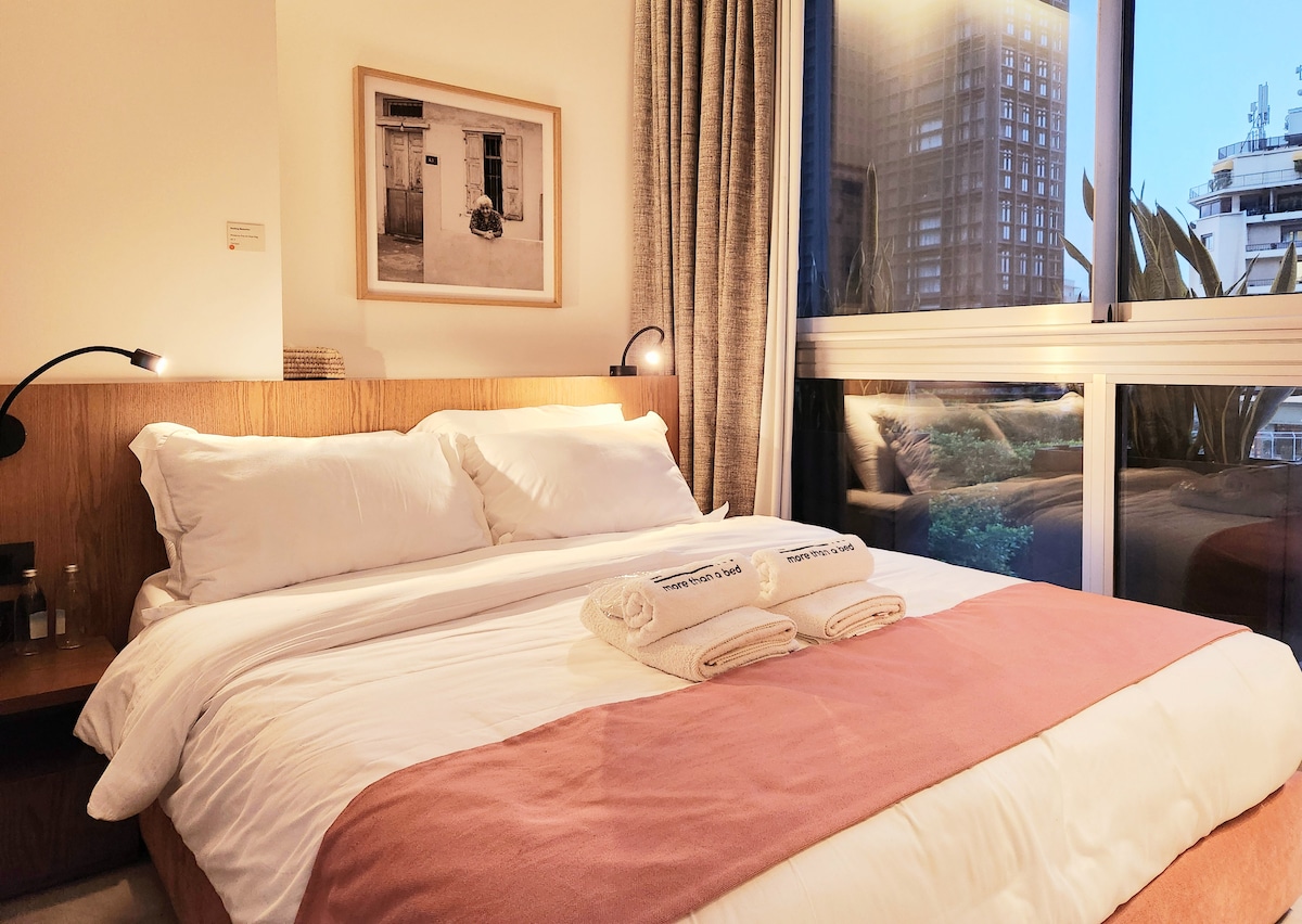 A modern bedroom features a spacious bed with crisp white linens and minimalist decor. A soft pink blanket is neatly laid at the foot, accompanied by two folded towels. Natural light flows through large windows, reflecting the urban landscape outside.
