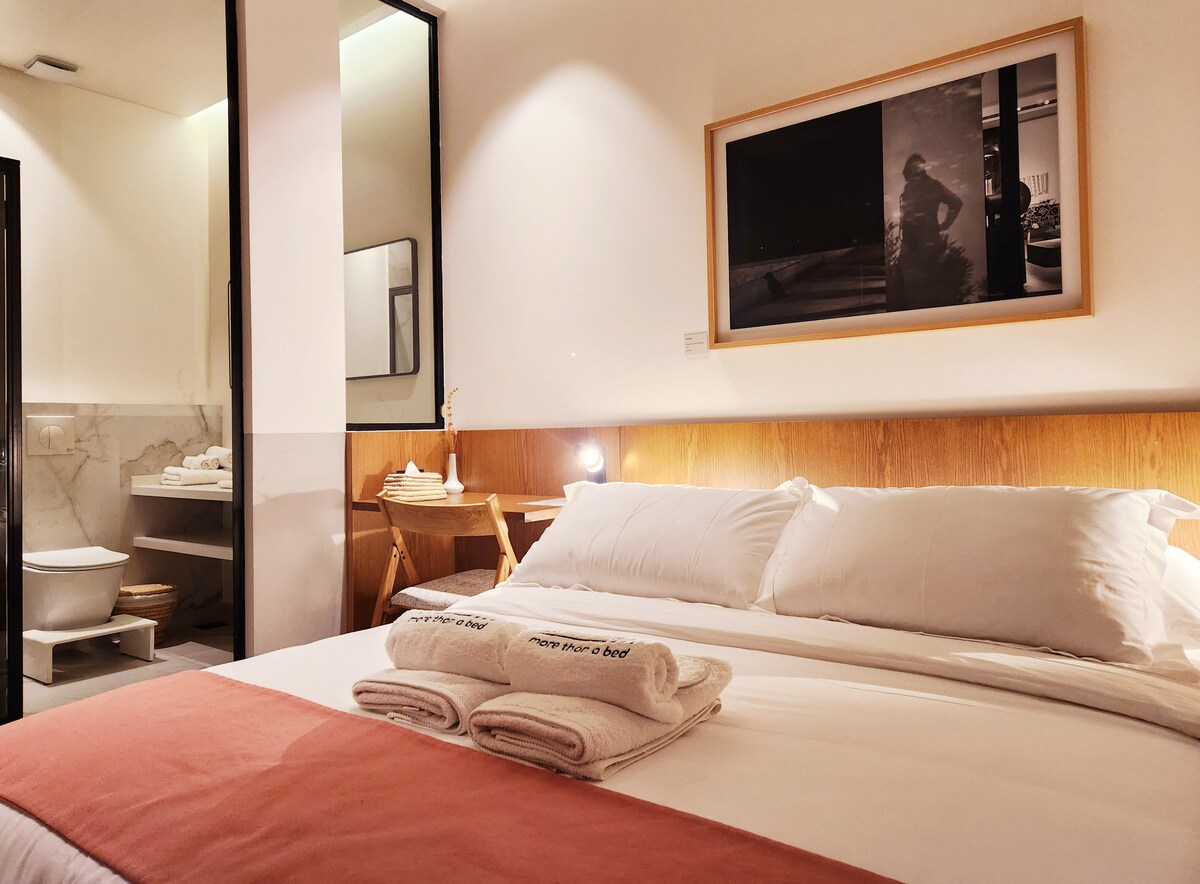 A contemporary room features a spacious bed with neatly folded towels at the foot. A softly lit area showcases a wooden frame, a large artwork on the wall, and open access to a modern bathroom designed with clean lines.
