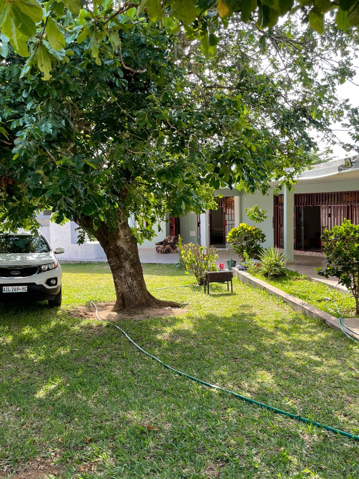 A spacious yard features a green lawn dotted with trees, providing shade and a natural ambiance. A parked vehicle is visible nearby, while a small table stands on a patio surrounded by well-kept shrubs and plants. The house is partially seen in the background.