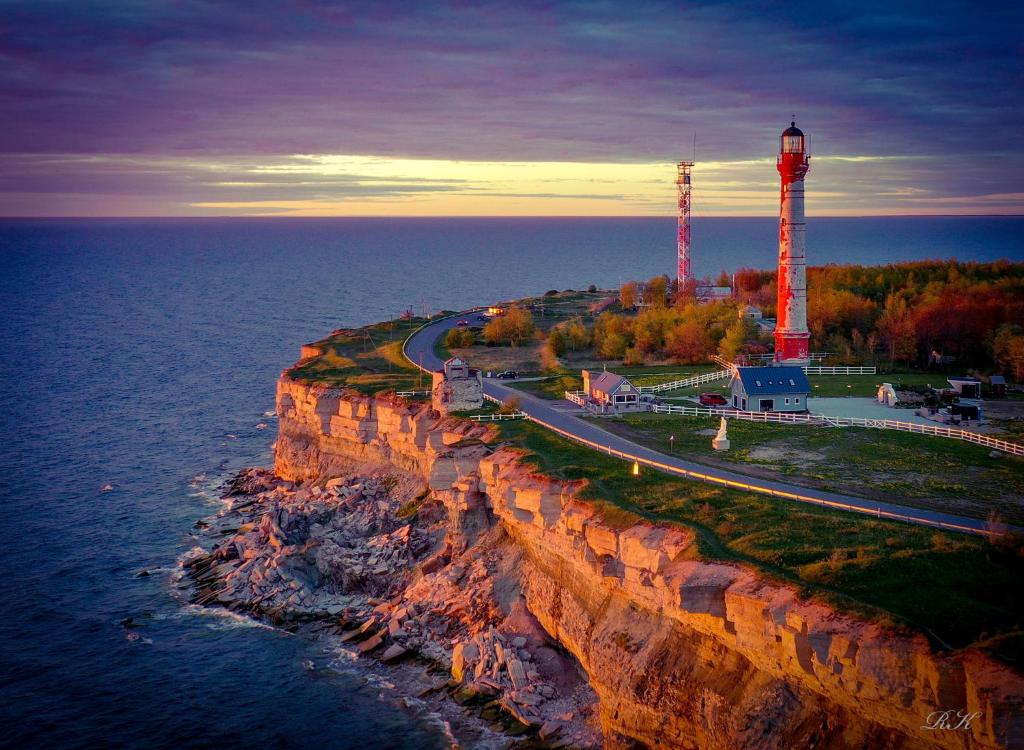 The image depicts a coastline featuring layered cliffs and a scenic road. Two lighthouses stand prominently, with one tall and red, situated near greenery. The sky transitions from purple to orange near the horizon, while the Baltic Sea reflects the soft hues of early evening light.