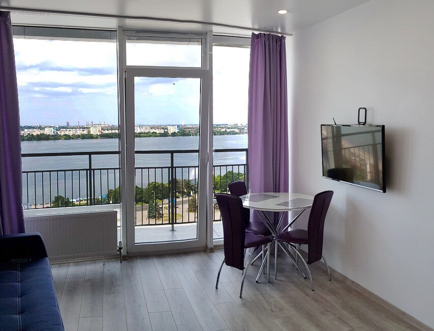 A modern living space features large glass doors that provide a waterfront view. A round table with purple chairs is positioned near the window, and a flat-screen TV is mounted on the wall. Light-colored flooring and purple curtains enhance the room's brightness.