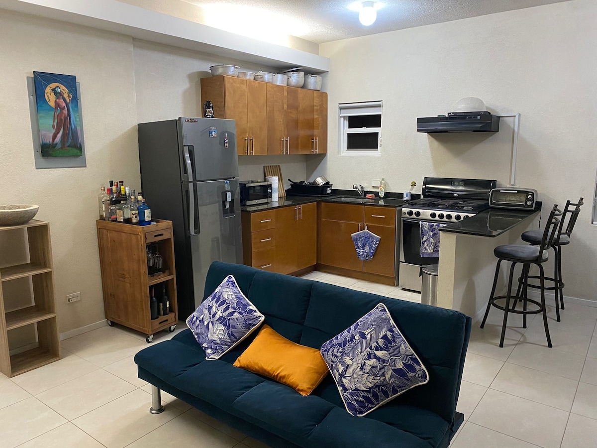 An open kitchen area features modern appliances, including a stainless steel refrigerator and a four-burner stove. The stylish counter is accompanied by bar stools. A comfortable blue sofa with decorative pillows is positioned in the inviting living space, while wall art adds a touch of character.