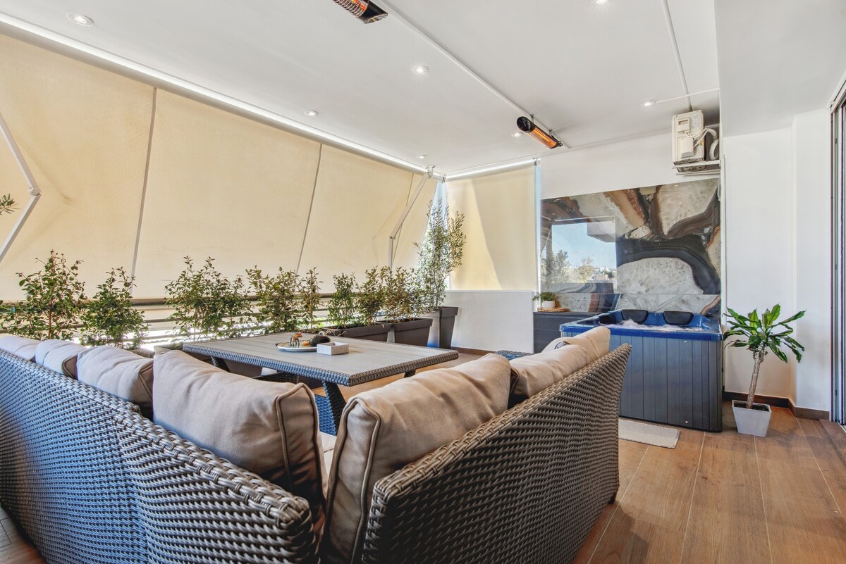 A spacious veranda is showcased with comfortable seating arranged around a sizable table. Lush greenery lines the edges, offering a serene atmosphere. A private Jacuzzi can be seen against a reflective wall, enhancing the outdoor experience.