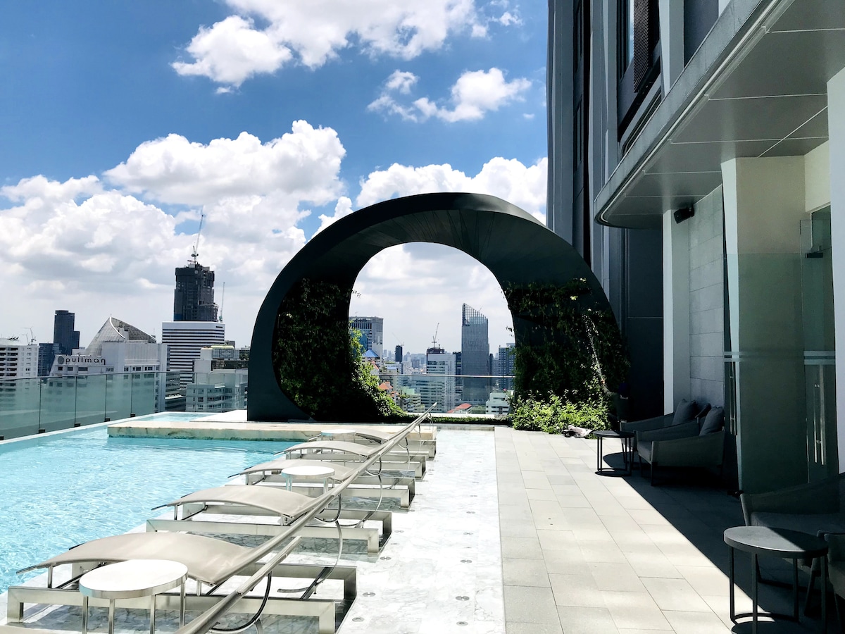 An outdoor terrace features a swimming pool with a city view. Lounge chairs are arranged along the pool's edge, complemented by a curved archway covered in greenery. The backdrop displays a skyline with various buildings under a bright sky with scattered clouds.