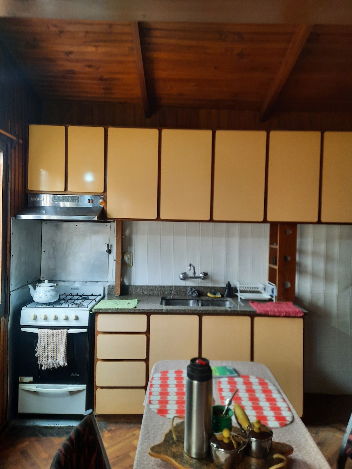 A functional kitchen is equipped with a gas stove, oven, and ample cabinetry. Counter space is neatly arranged with kitchen essentials, including a kettle and utensils. A checkered tablecloth covers a dining table, complemented by wooden accents and a rustic ceiling featuring exposed beams.