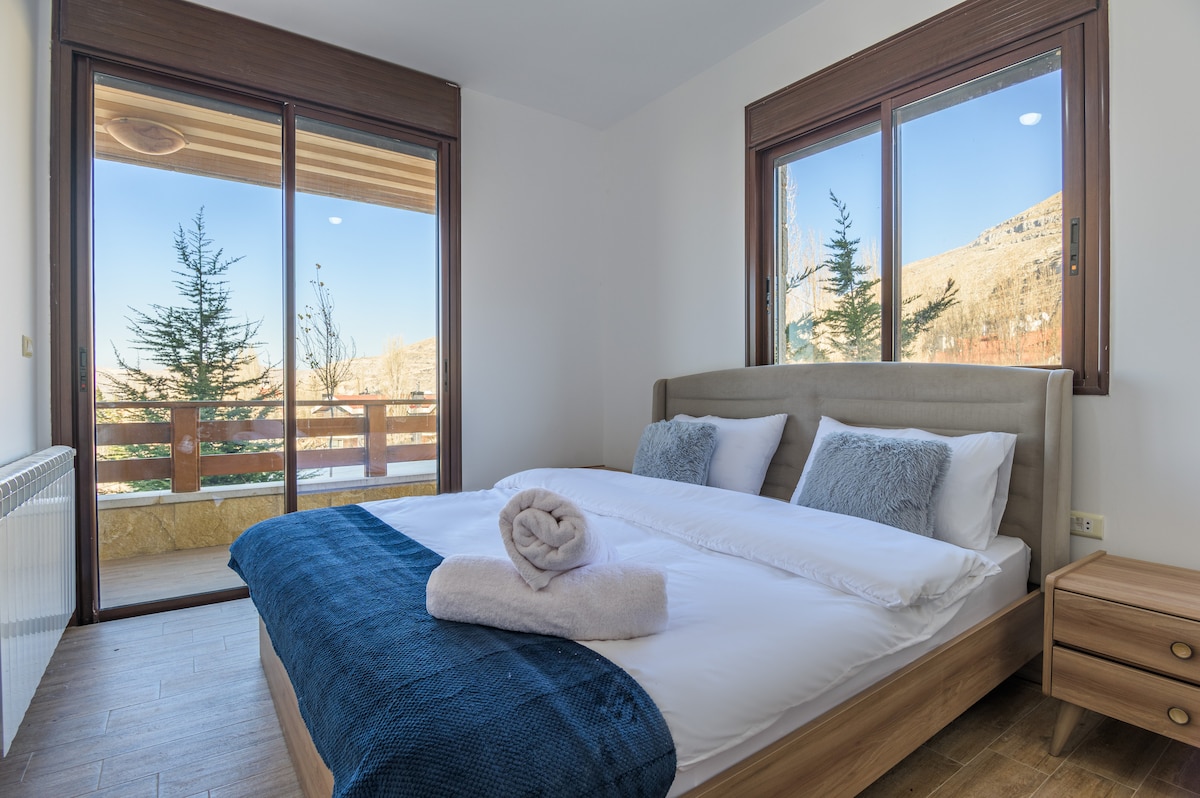 A cozy bedroom is highlighted by a spacious bed dressed in crisp white linens and a deep blue throw blanket. Large windows provide natural light and views of the outdoors, while a balcony can be seen through the glass doors.