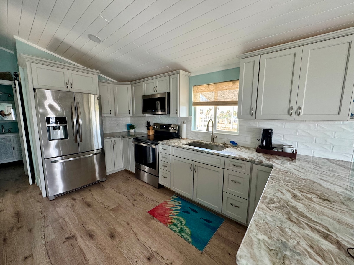 A modern kitchen features stainless steel appliances, including a refrigerator and stove. Light grey cabinetry complements the marble countertop. A coffee maker and cookware are available, while a colorful rug adds a touch of warmth to the space. Natural light filters through the window.