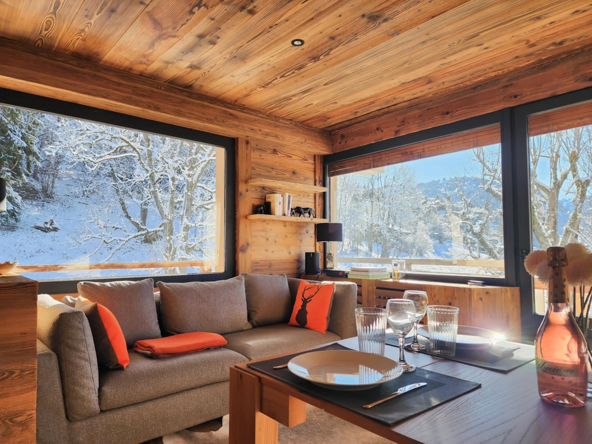 The image showcases a cozy living area with a plush sofa adorned with orange accent cushions. Large windows reveal a snowy landscape, allowing natural light to fill the space. A wooden dining table is set with glassware, enhancing the inviting atmosphere.
