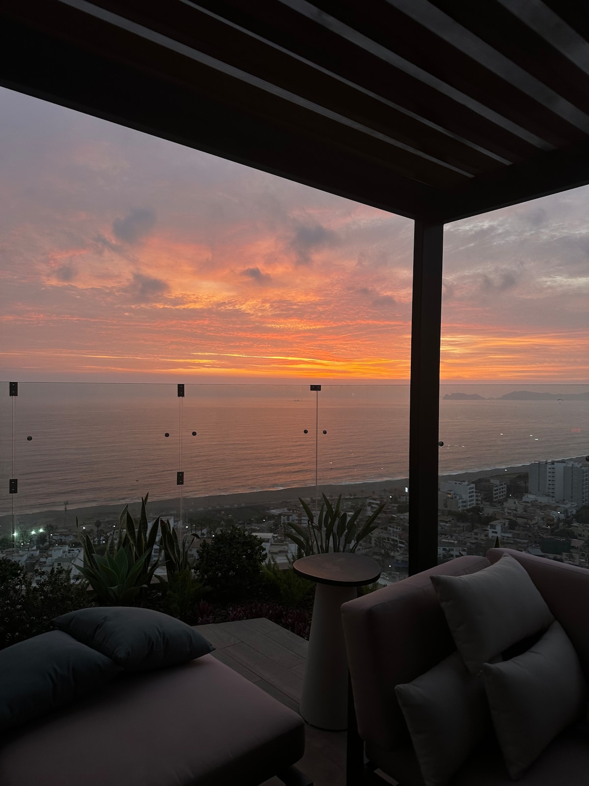 A private balcony offers sweeping views of a vibrant sunset over the ocean. The tranquil scene is framed by a modern glass railing and lush greenery, creating a serene space for relaxation. Plush seating provides comfort while taking in the breathtaking panorama.