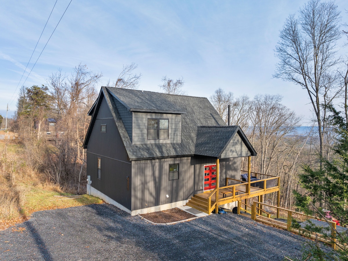 The exterior of a modern mountain home is presented, featuring a sleek gray facade and a bold red front door. A wooden deck extends from the entrance, surrounded by trees and a gravel driveway, suggesting a serene setting in a natural landscape.