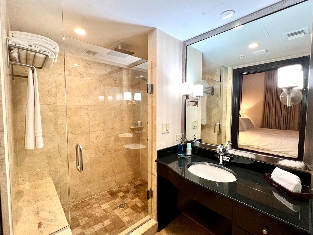 The bathroom features a walk-in shower with natural stone walls and a glass enclosure. A sleek, dark vanity with a round sink is accompanied by stylish lighting fixtures. A large mirror reflects the sleeping area, while fresh towels hang on a nearby rack.