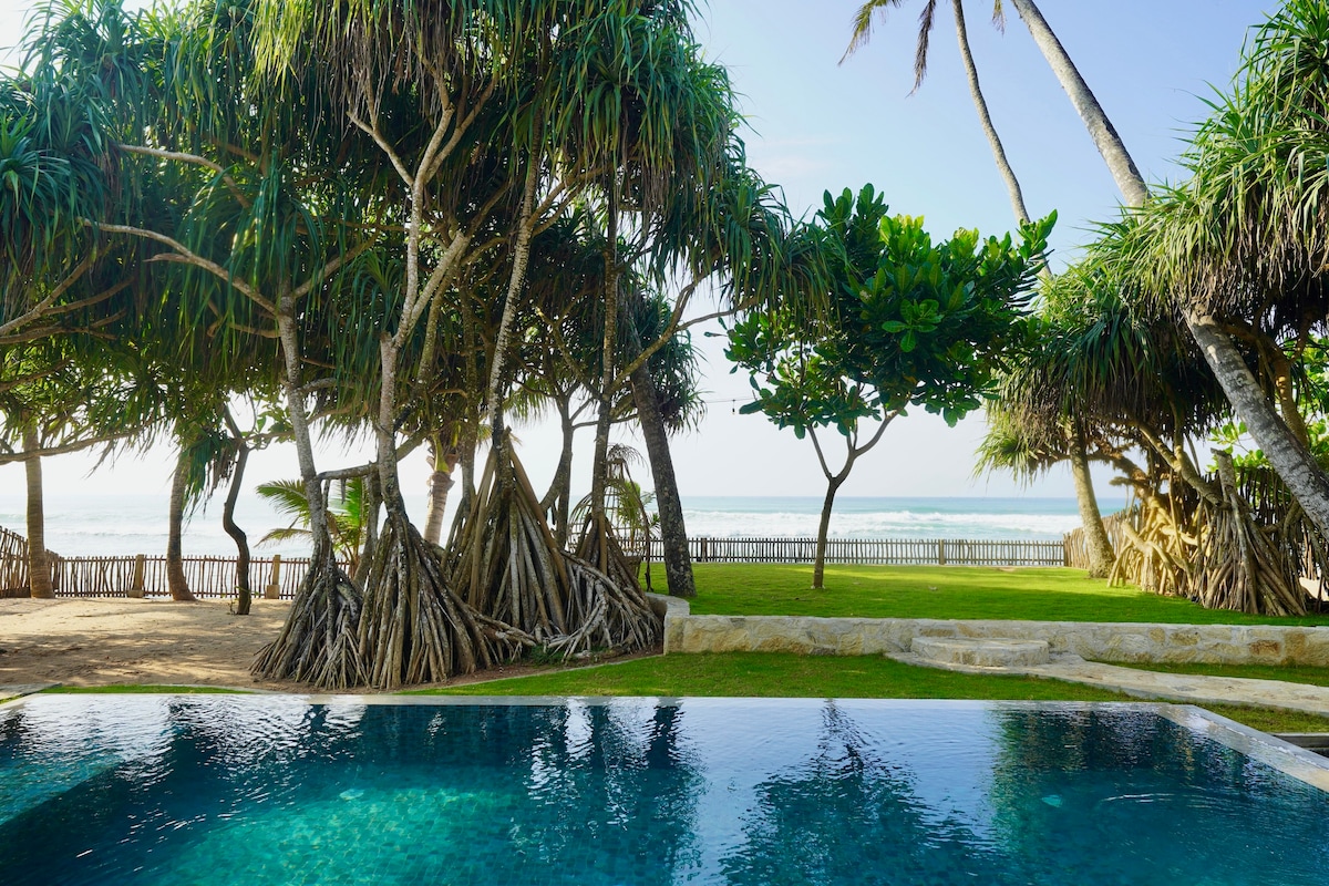A crystal blue pool reflects the sky, surrounded by lush gardens and tall pandanus trees. The ocean is visible in the background, creating a serene coastal atmosphere. Gentle waves can be seen meeting the sandy shore.