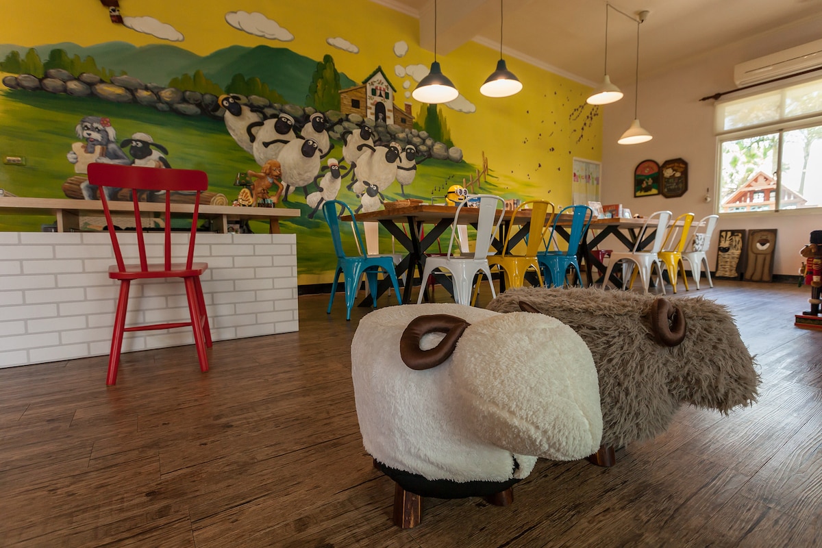 A lively dining area is highlighted by a colorful mural depicting sheep in a pastoral setting. Various dining tables with mixed seating of colorful chairs complement the playful atmosphere. Unique sheep-shaped stools add a charming touch to the space, inviting relaxation and enjoyment.
