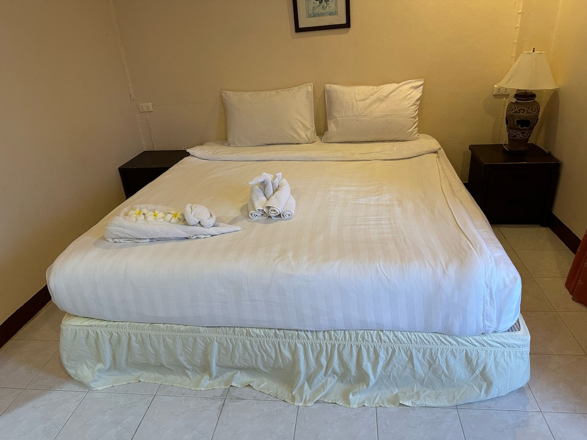 A spacious bed is centered in the room, dressed in crisp white linens. Neatly arranged towels are placed on the bed, flanking a decorative lamp on a nightstand. Soft lighting adds a warm touch to the neutral-colored walls.