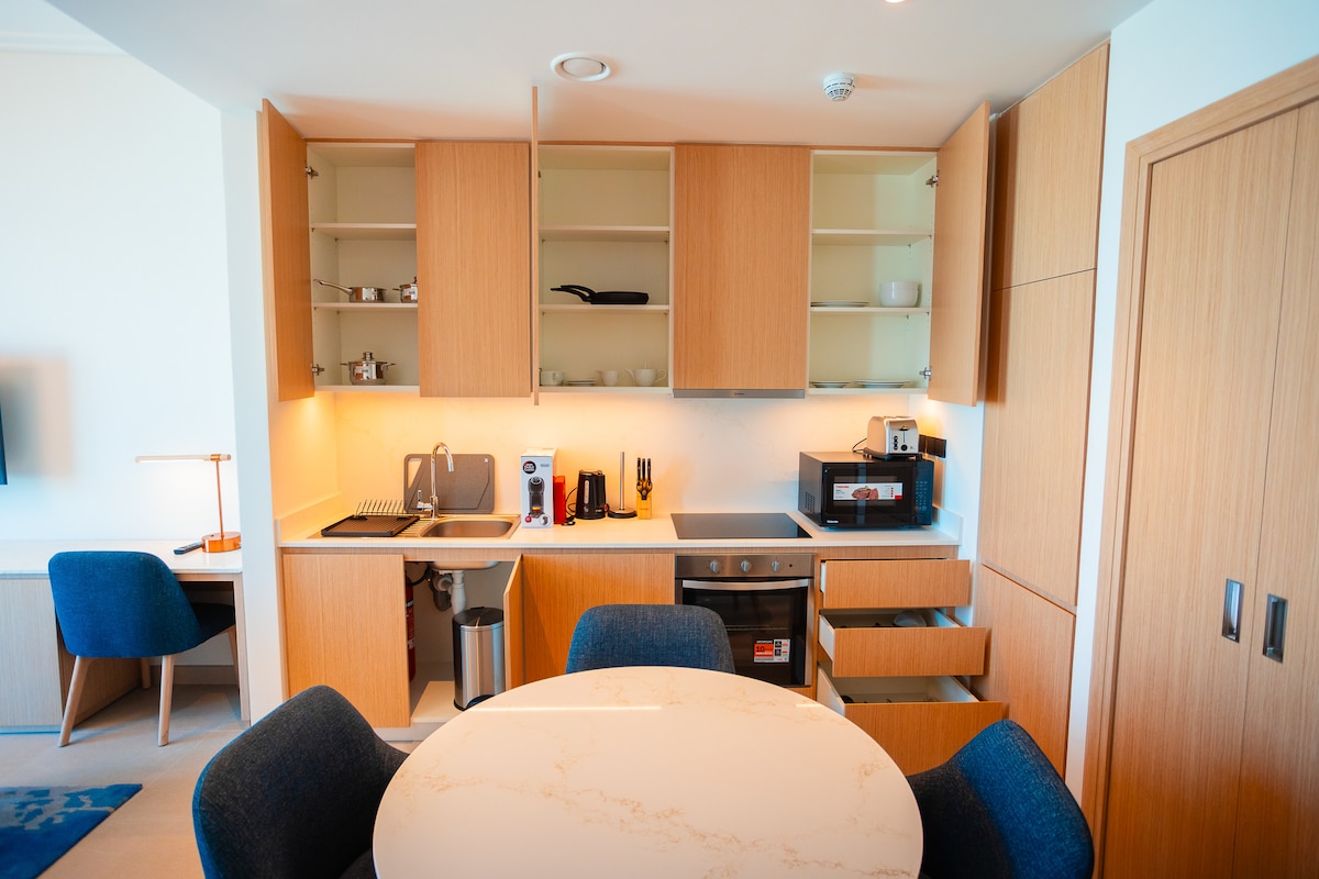 A modern kitchen area is shown with wooden cabinetry and a smooth countertop. Appliances include a coffee maker, microwave, and an oven. A round dining table with four cushioned chairs is positioned nearby, creating a welcoming dining space.