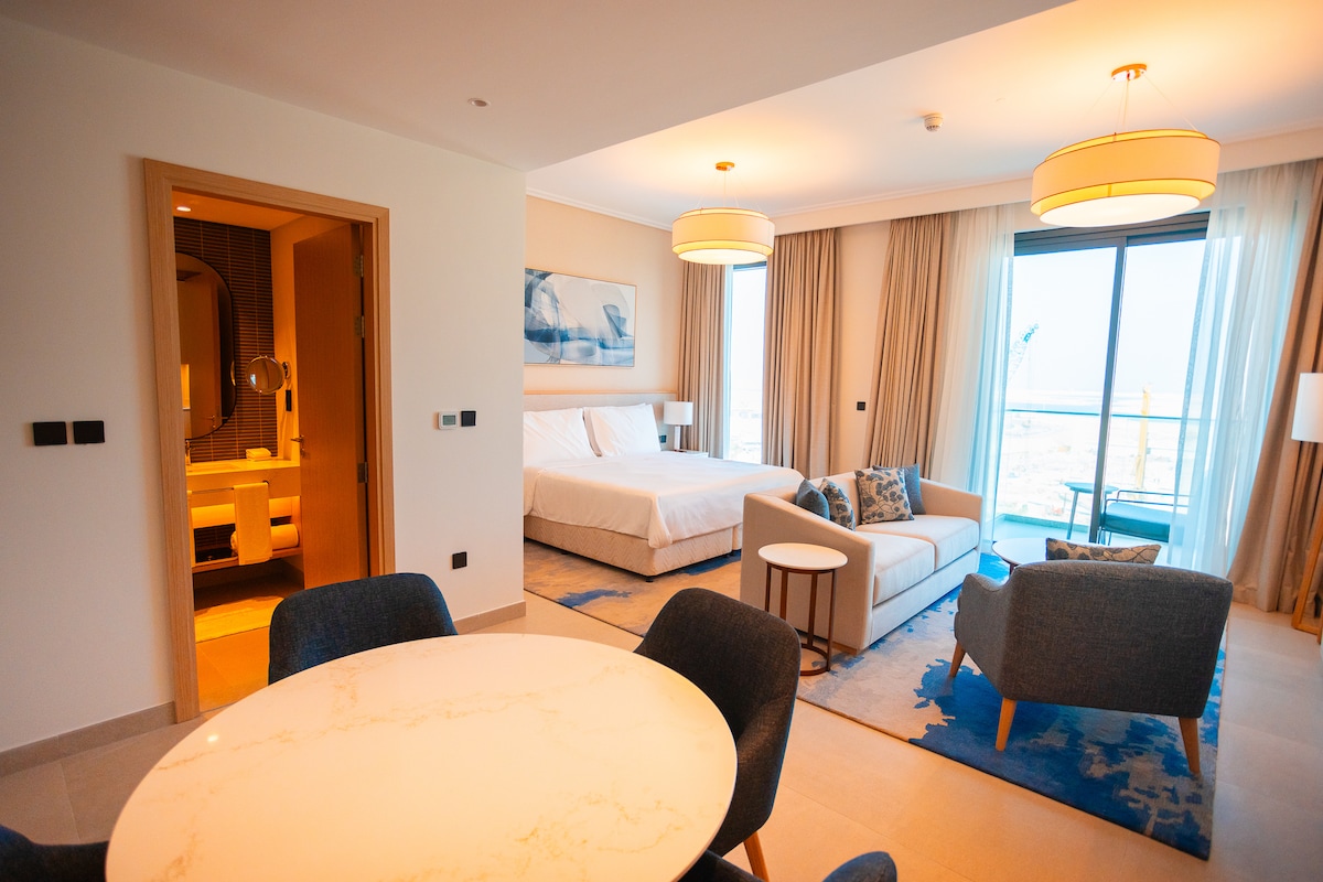 An open studio layout is showcased, featuring a spacious bed with a light headboard and two matching armchairs. A round dining table sits nearby, and an expansive window allows natural light to fill the space, offering views of the surroundings.