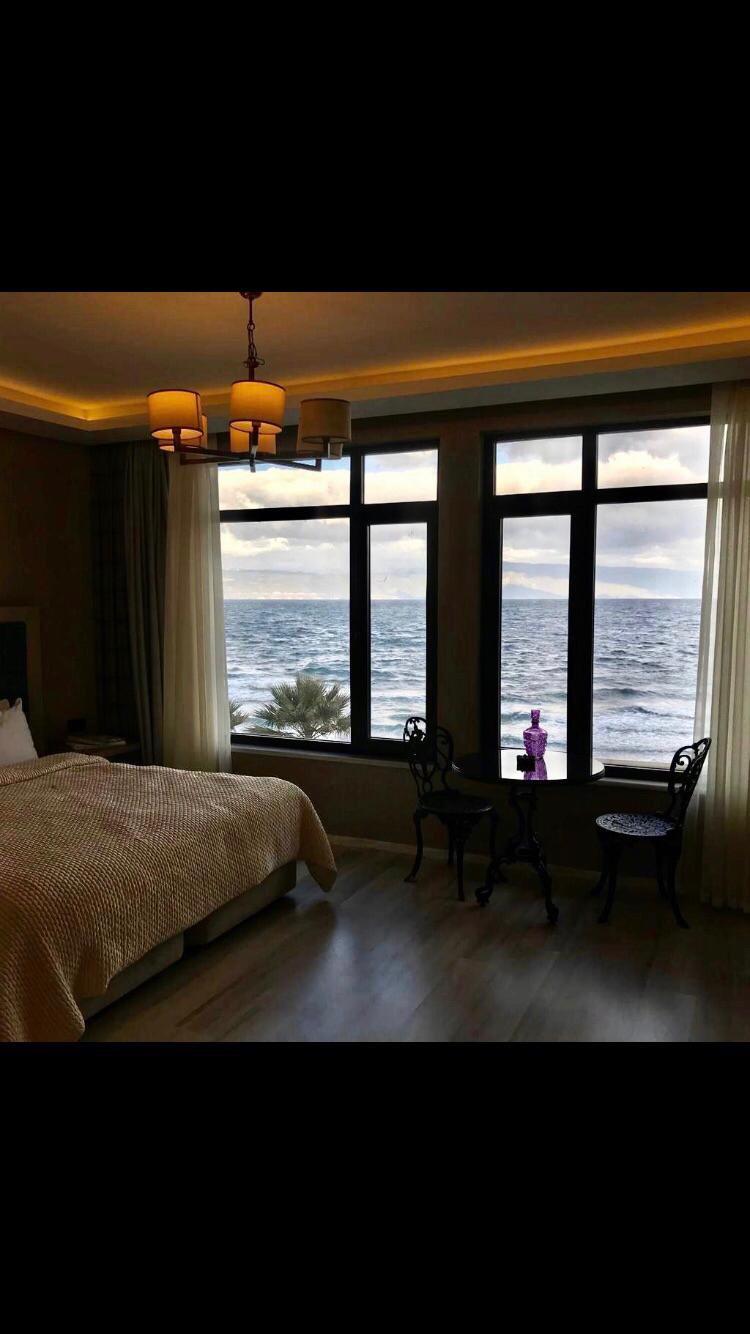 A serene bedroom features large windows with views of the sea, letting in natural light. The space includes a spacious bed with a textured throw, and two elegant chairs positioned near the windows, providing a cozy spot to enjoy the view.