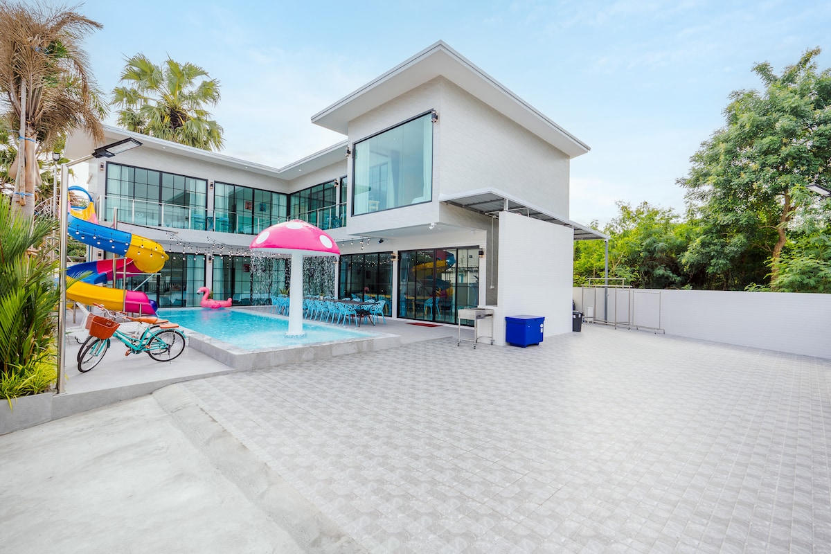 An outdoor area is displayed featuring a large swimming pool surrounded by a spacious patio. Colorful water slides are visible next to the pool, along with a pink mushroom-shaped feature. Lush greenery borders the scene, enhancing the inviting outdoor space.