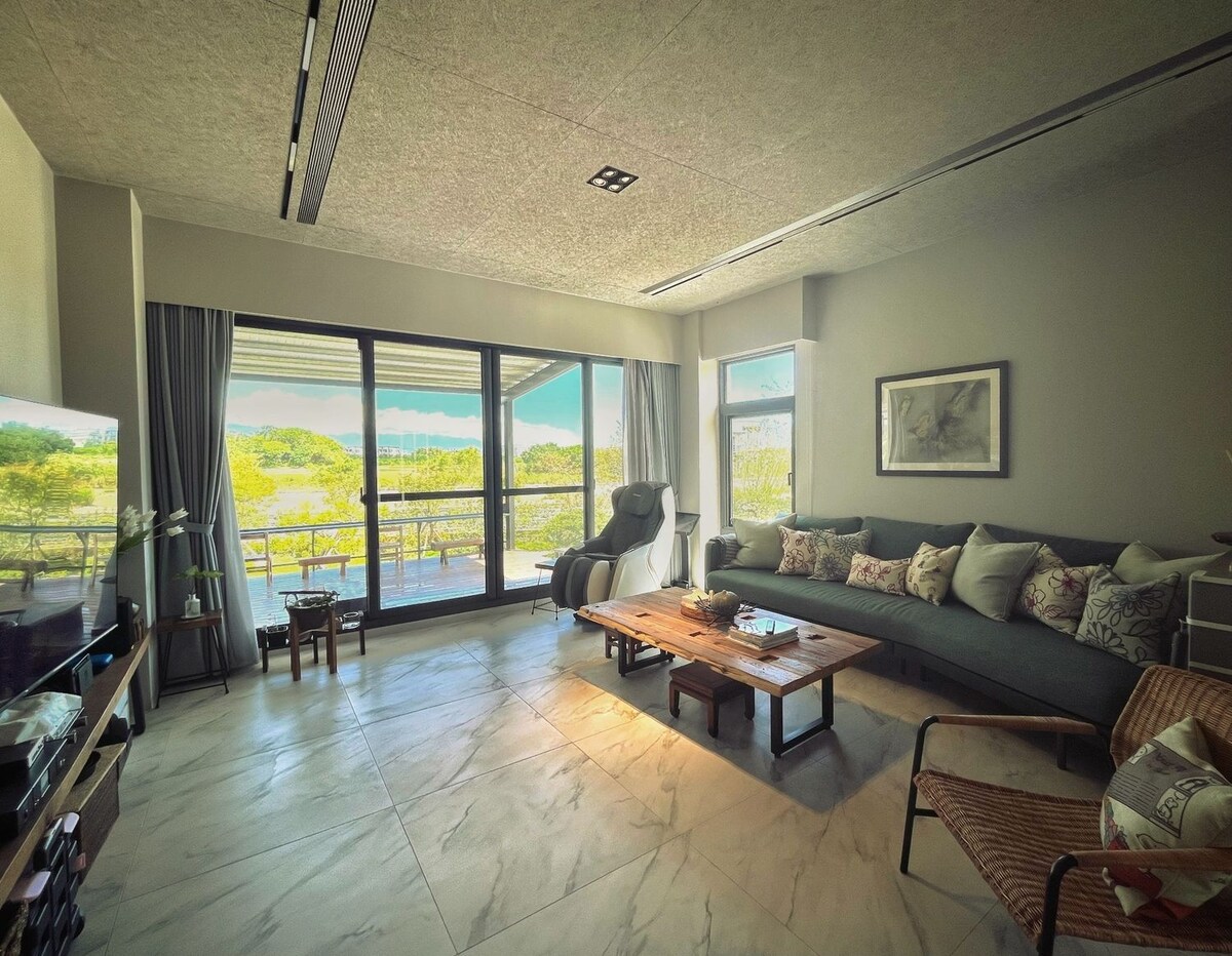 A spacious living room features large glass doors inviting natural light. A comfortable sectional sofa is positioned alongside a coffee table, while a recliner offers additional seating. The backdrop includes a view of greenery, enhancing a serene ambiance.