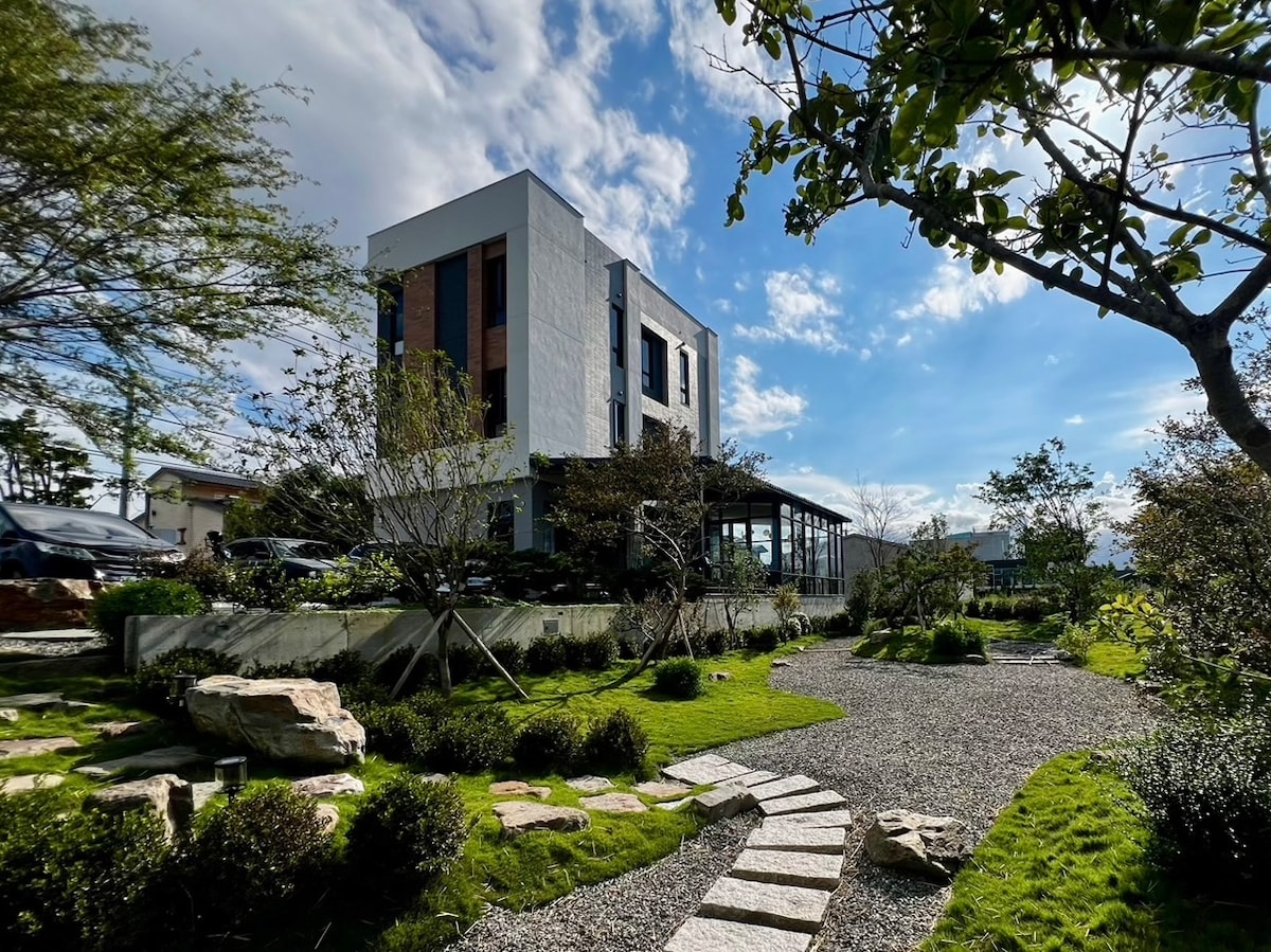 A modern building with a white stone facade is surrounded by a landscaped garden. A stone pathway leads through the lush greenery, with bushes and small trees dotting the area. The sky is partly cloudy, creating a serene outdoor atmosphere.