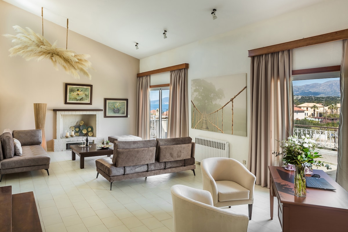 A spacious living room features cozy seating arranged around a central coffee table. Large windows offer a view of the surrounding landscape, with soft curtains framing the natural light. A modern art piece and decorative accents enhance the elegant atmosphere.