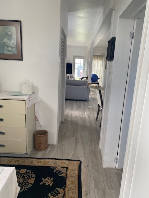 A well-lit hallway connects different areas of the cottage, featuring light-colored walls and tiled flooring. A dresser stands on the left, accompanied by a decorative rug. A living area is visible in the distance, with natural light filtering through nearby curtains.