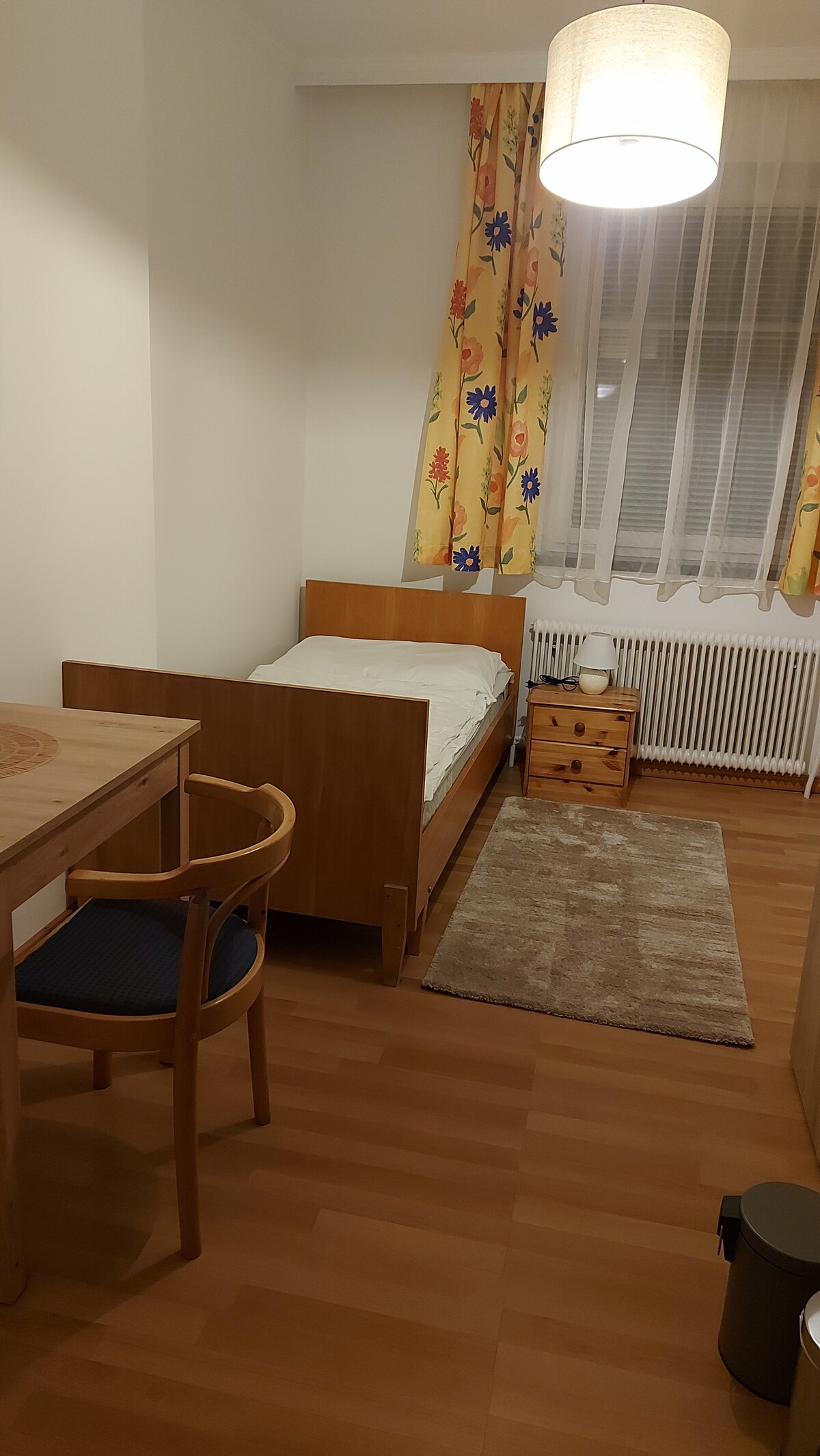 A simple bedroom features a single bed with a light-colored mattress, accompanied by a wooden bedside table and a small lamp. A wooden desk with a chair is positioned beside a window adorned with colorful curtains, which let in soft natural light.