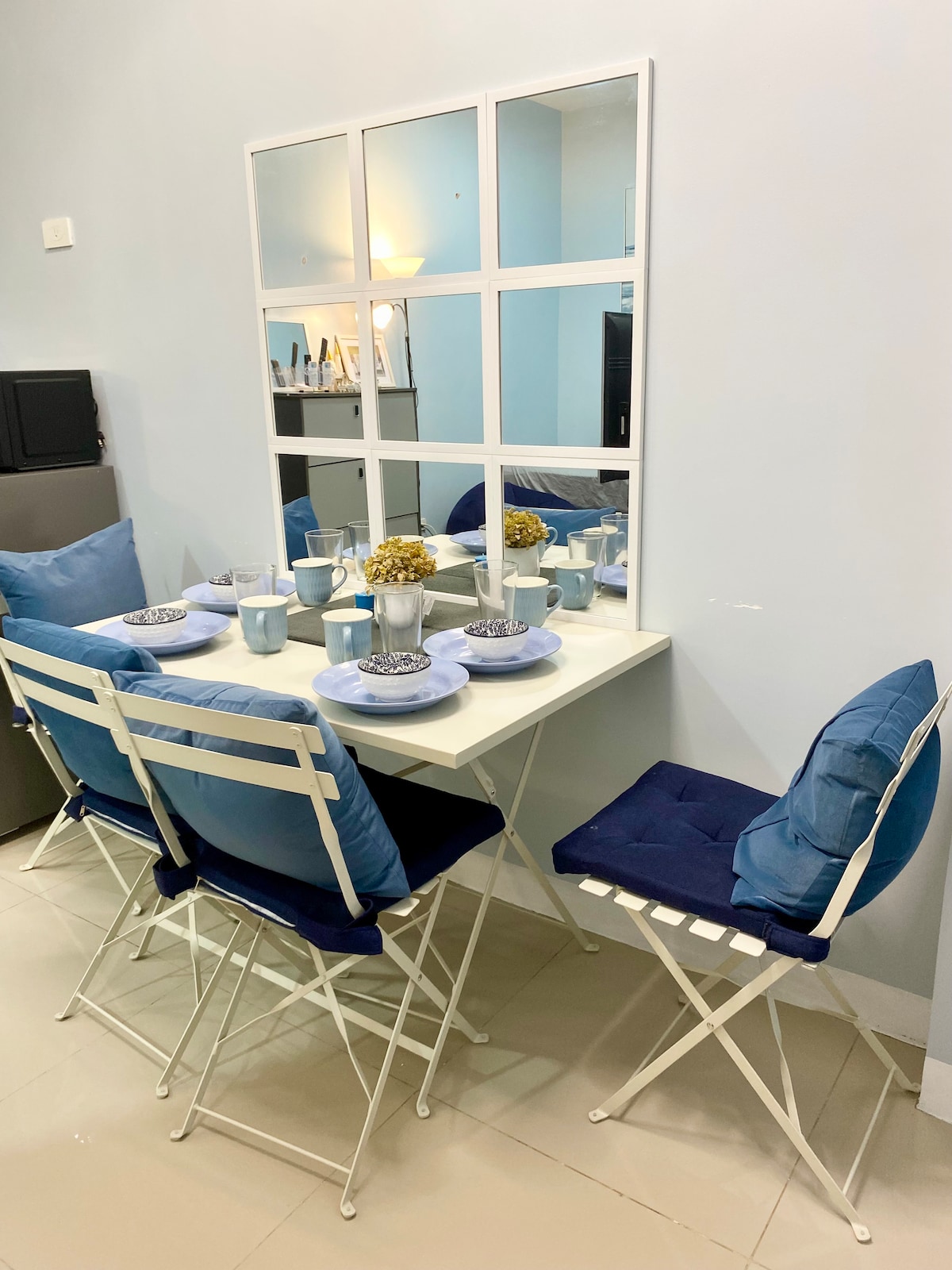 A dining area features a white table surrounded by four blue cushioned folding chairs. A decorative mirror with a grid design reflects the space, while the table is set with blue and silver tableware, creating an inviting setting for meals.