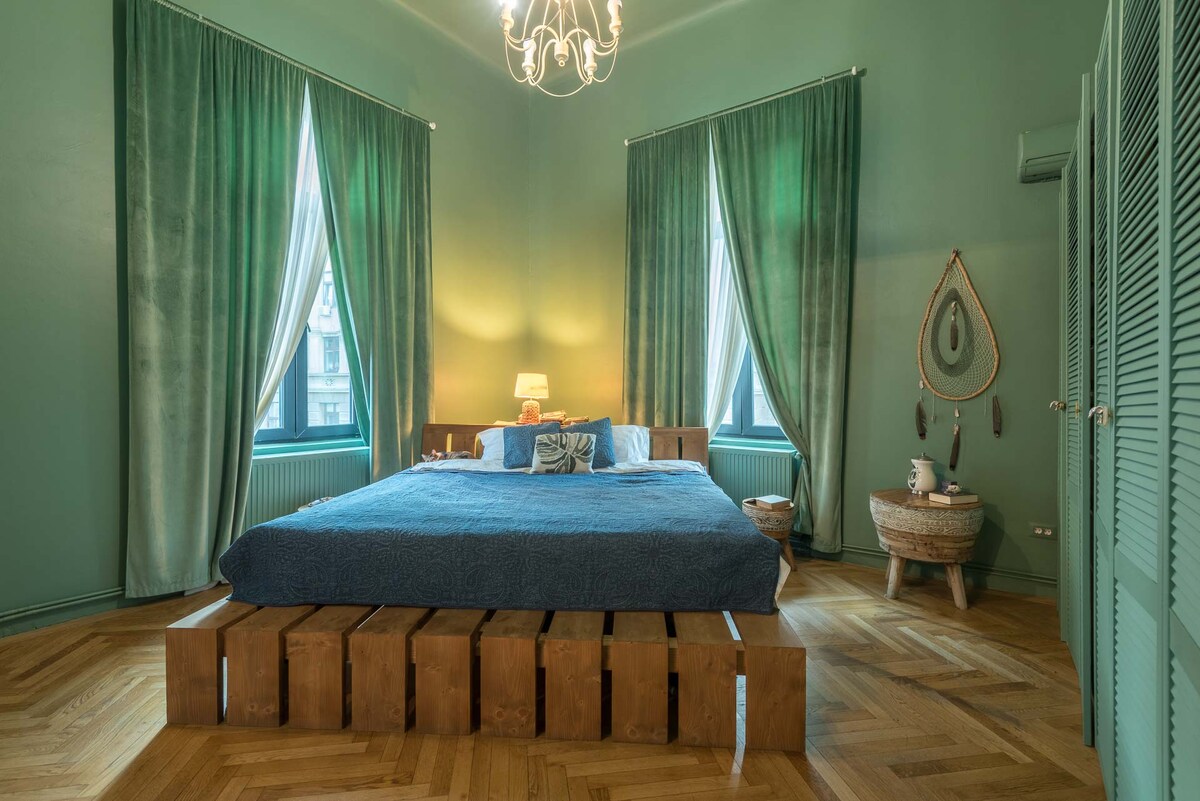 A calm bedroom features a large bed with a blue coverlet, surrounded by floor-to-ceiling curtains in coordinating green. Two windows allow natural light to fill the space, while a wooden accent element adds warmth. Decorative items are arranged tastefully on a side table.