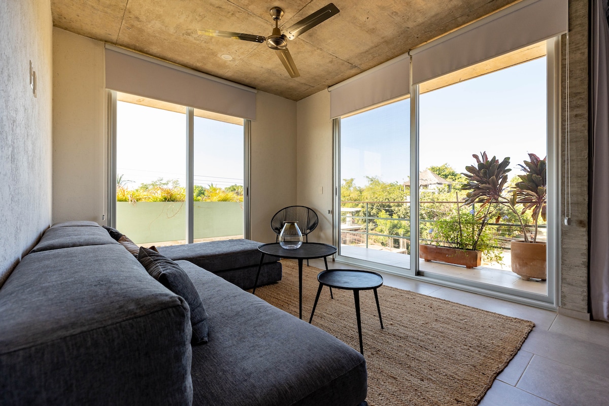 A modern living area features large windows that frame a view of lush greenery. A sleek gray sectional couch complements a minimalist black coffee table. A round rug adds warmth to the space, while a ceiling fan circulates air, enhancing the relaxed ambiance.