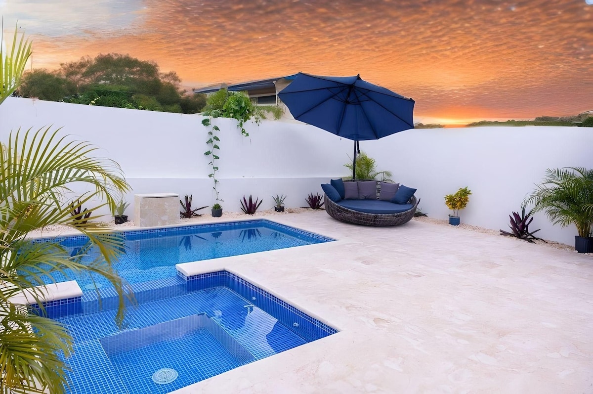 A private outdoor area showcases a sparkling blue swimming pool adjacent to a relaxing jacuzzi. A shaded seating area is provided by a large umbrella, complemented by surrounding tropical plants and flowers. The scene is set against a vibrant sunset sky.