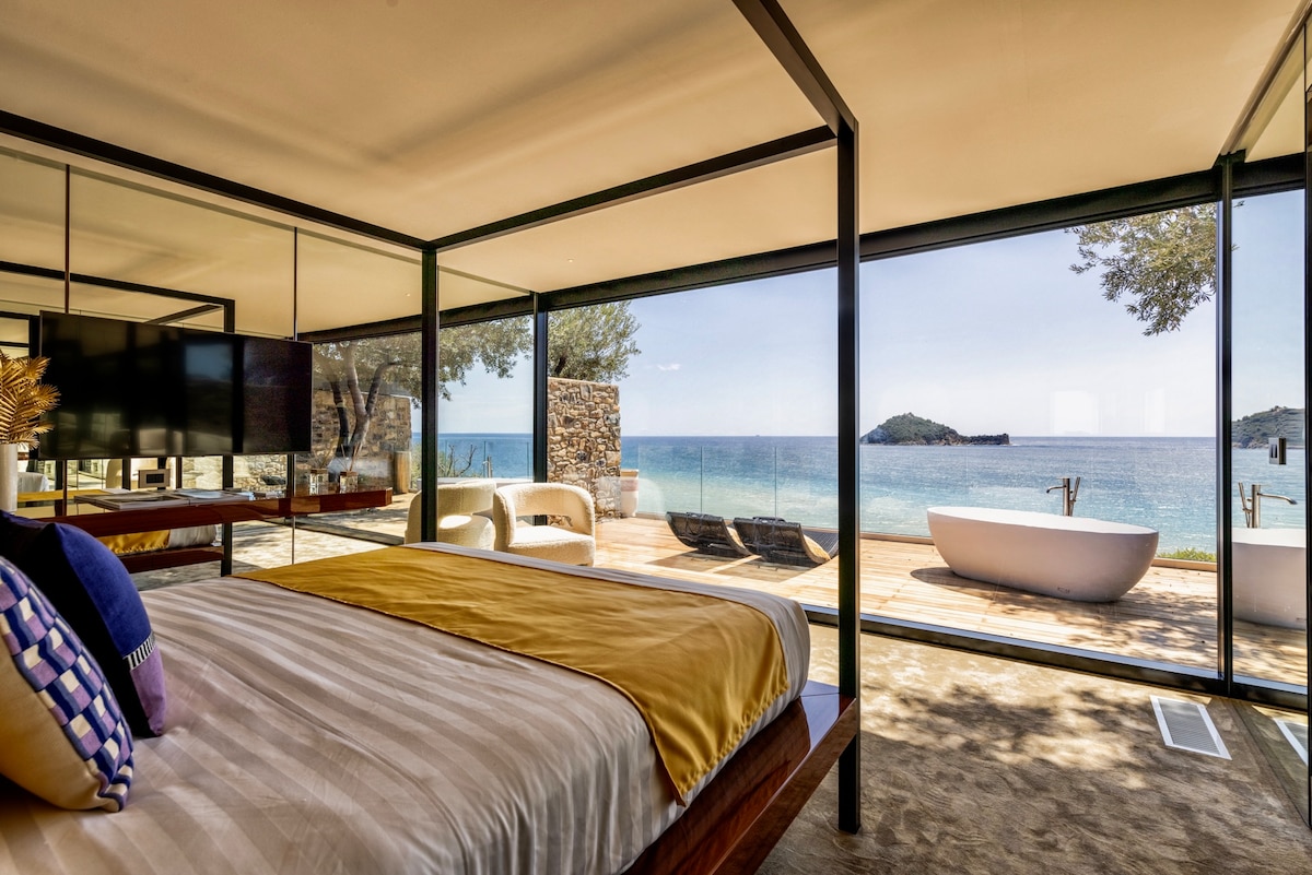 A spacious suite features a large bed facing expansive glass walls that offer stunning views of the sea and island. A seating area with modern furniture is visible on the terrace, along with a freestanding bathtub. Natural light fills the room, creating an inviting ambiance.
