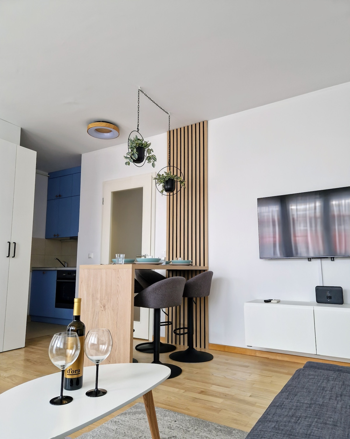 A modern living area showcases a light wood table set with two wine glasses and a bottle of wine. Two bar stools accompany a small kitchen island. A television is mounted on the wall, and hanging plants add a touch of greenery.