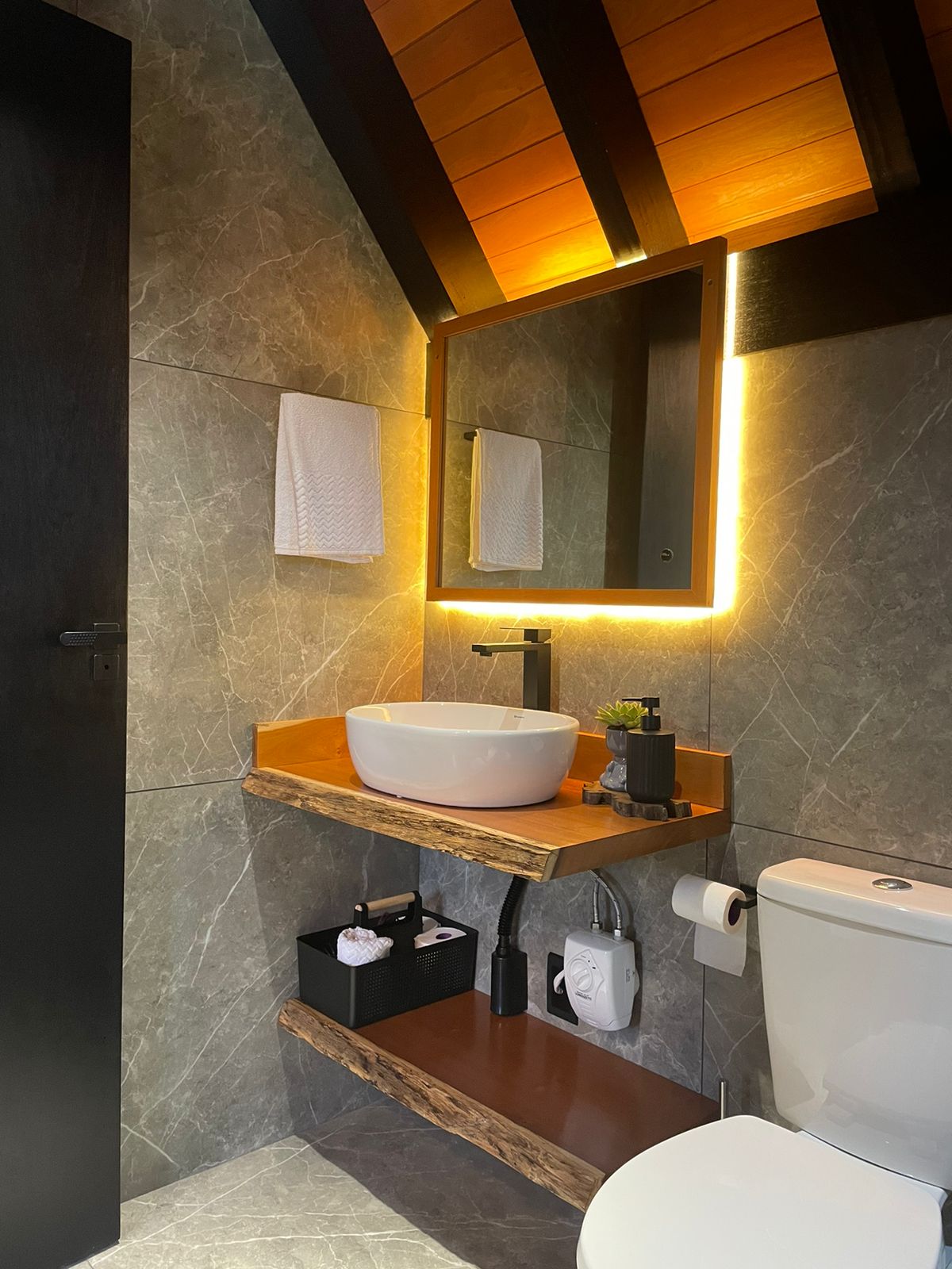 The bathroom is highlighted by natural stone walls and wooden accents. A contemporary round sink is placed atop a wooden shelf, while a mirror with ambient lighting enhances the space. Towels and accessories are neatly arranged, contributing to a clean and functional environment.