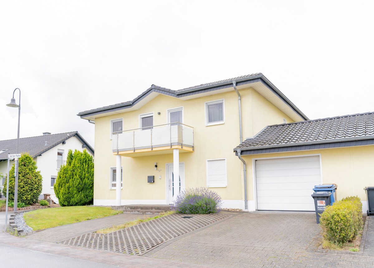 The exterior of a two-story house is presented, featuring a light yellow facade and a balcony above the entrance. A driveway leads to a garage on the right, while neat landscaping includes green shrubs and a flowering plant near the entrance.