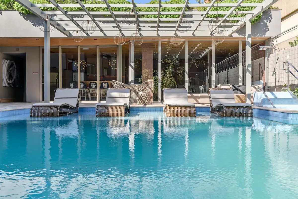 A serene pool area features crystal-clear water surrounded by comfortable lounge chairs. A shaded overhead structure offers a blend of indoor and outdoor seating, enhancing the inviting ambiance. Stylish decor is visible in the background, creating a relaxed atmosphere for guests.