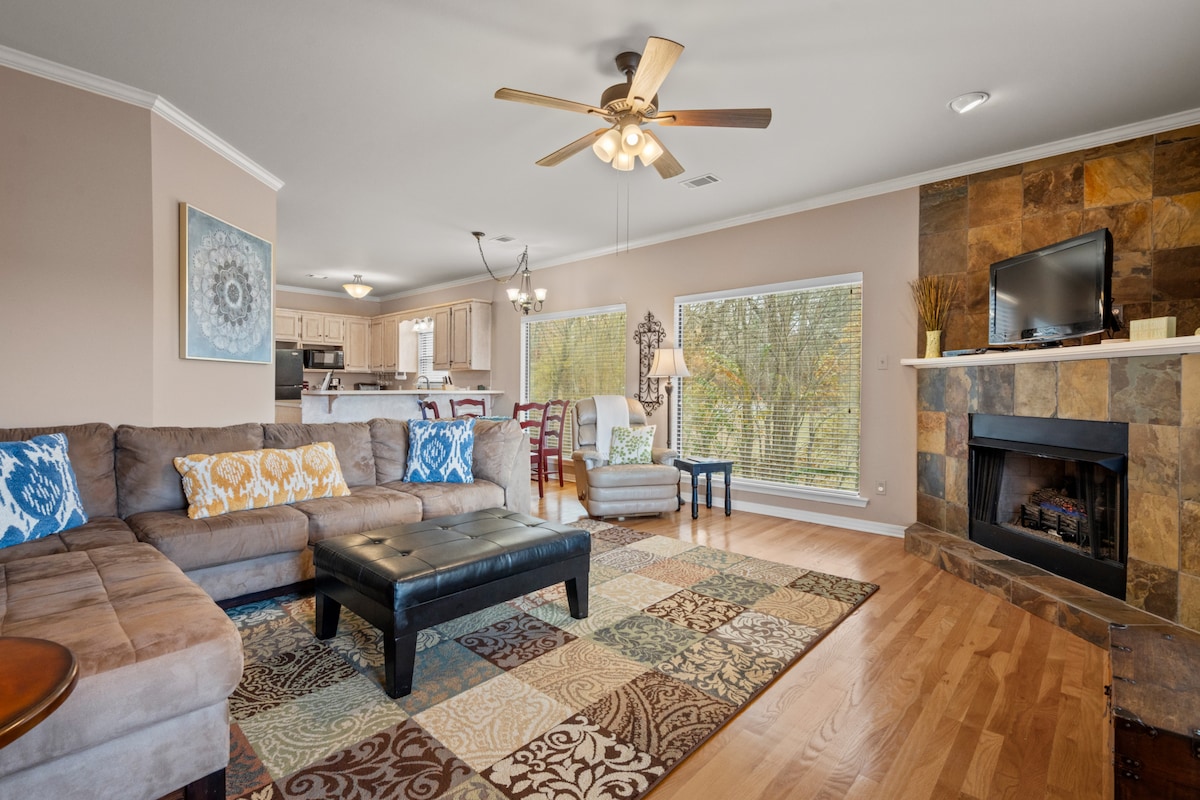 A spacious living area features a large sectional sofa adorned with decorative cushions and a patterned area rug. Natural light fills the space through expansive windows, and a flat-screen TV is mounted above a stone fireplace, creating a welcoming ambiance for relaxation.