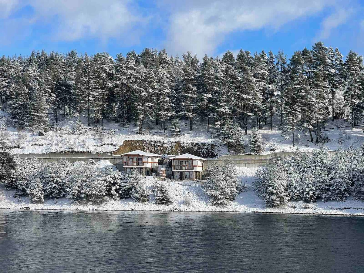 A view of two modern buildings set against a snowy landscape, reflected in a calm lake. Tall trees with a dusting of snow line the hillside, creating a serene and tranquil setting under a partly cloudy sky.