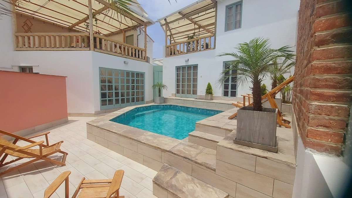 The courtyard features a refreshing pool surrounded by polished stone. Sun loungers are positioned near the poolside, and a tropical palm tree adds greenery. Large glass doors provide natural light to the adjacent areas, while a soft awning offers shade over the space.