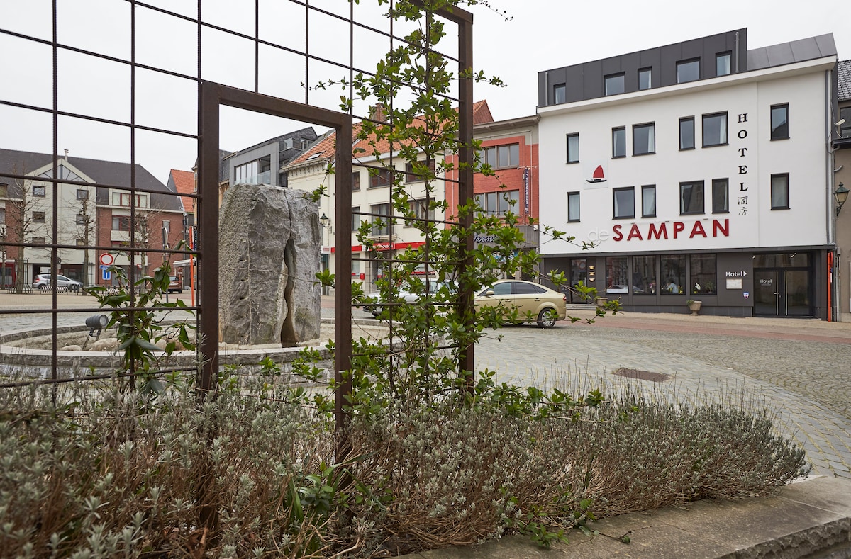 The image depicts a hotel building named 'Sampan' located in a central square. A tranquil square is framed by greenery and a stone sculpture, with urban architecture visible in the background. A vehicle is parked nearby, contributing to the accessibility of the location.