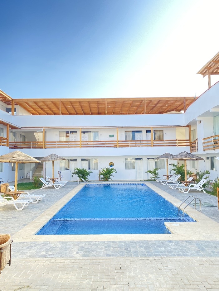 A central pool area is surrounded by lounge chairs and thatched umbrellas. The hotel structure features multiple levels with balconies overlooking the pool. Lush greenery adds a natural touch to the setting, creating a serene environment for relaxation.