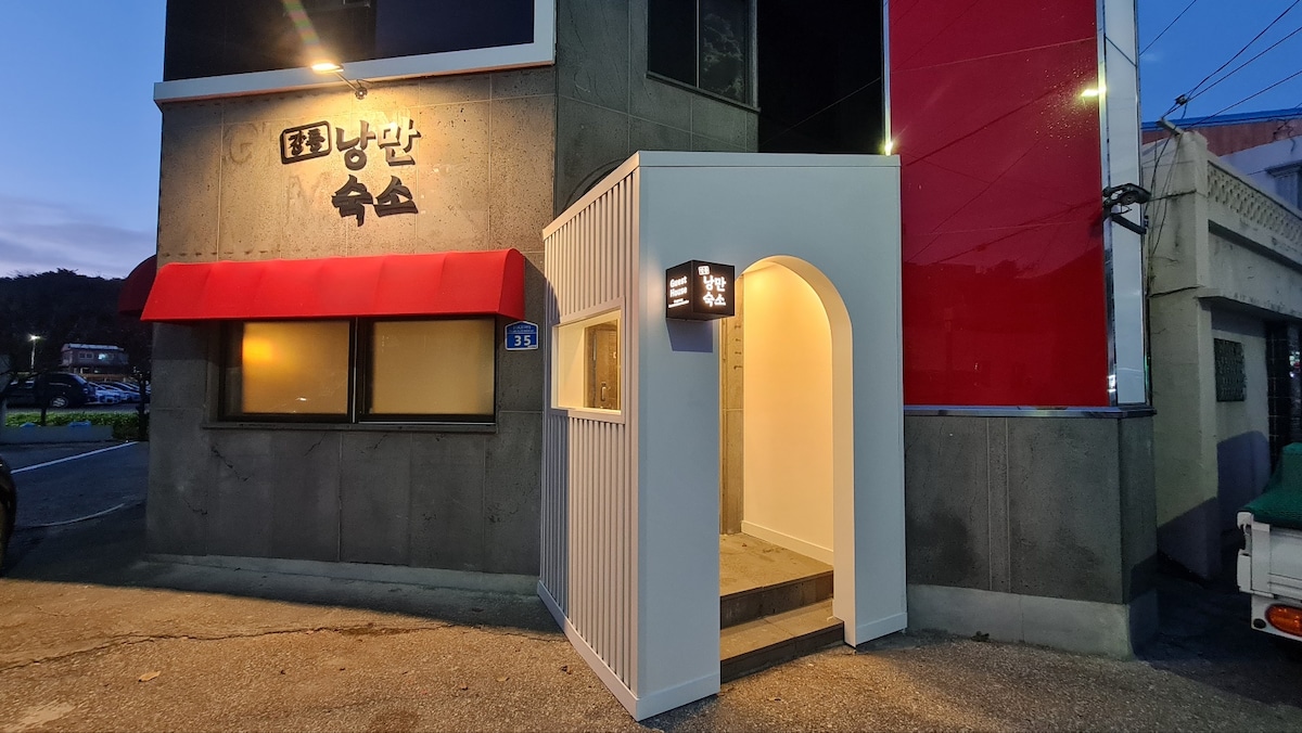 An entrance canopy leads to the building, showcasing a clean and modern design. The exterior features a combination of concrete and red accents, with warm lighting illuminating the doorway. A sign displays the property name, providing a welcoming presentation as dusk settles.
