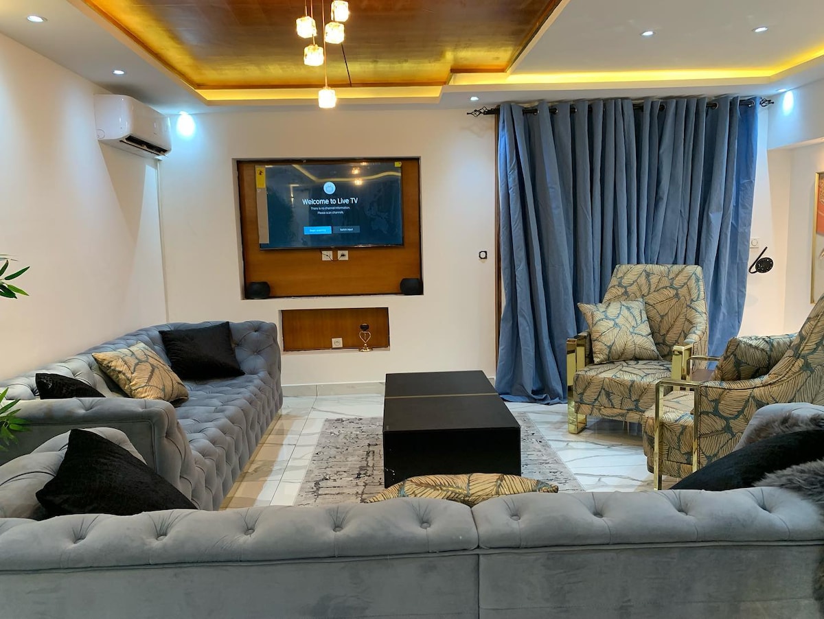 A spacious living room is depicted, featuring a plush gray sectional sofa with multiple cushions. A sleek coffee table is centered in the room, while a large flat-screen TV is mounted on the wall. Floor-to-ceiling dark curtains frame a stylish ambience.