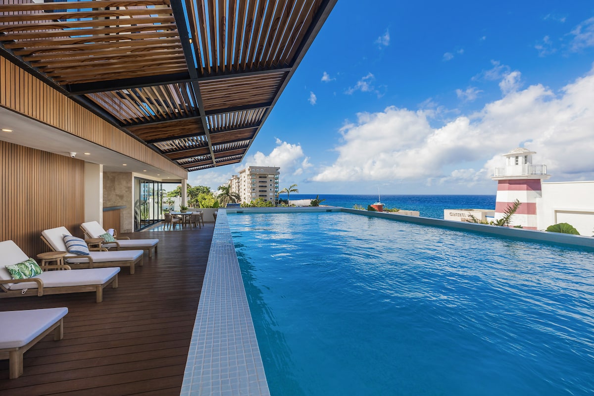 A serene infinity pool extends alongside a wooden deck, with several sun loungers arranged for relaxation. A panoramic view of the ocean and clear blue sky can be seen in the background, complemented by modern architectural elements.