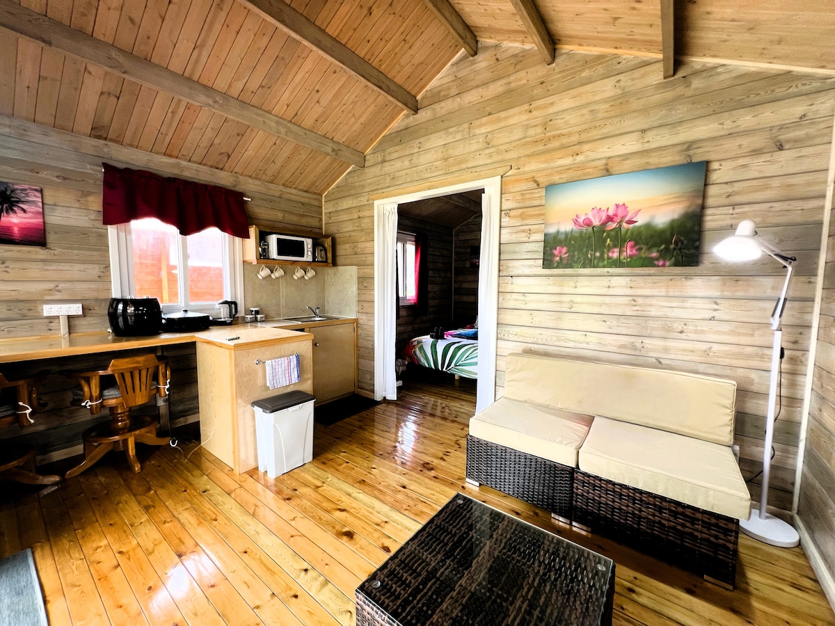 A cozy interior showcases wooden walls and ceiling beams, creating a warm atmosphere. A seating area with a neutral sofa is positioned beside a dark coffee table. An open kitchenette includes a microwave and a small dining table. A doorway leads to a bright bedroom with floral artwork.