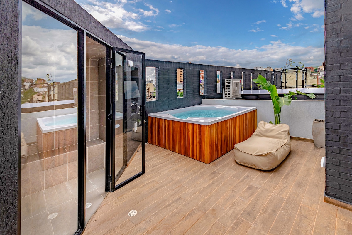 An outdoor terrace features a wood-paneled hot tub surrounded by a spacious wooden deck. Large glass doors provide access to the interior, while a potted plant adds a touch of greenery. The view includes cityscapes under a dramatic sky.