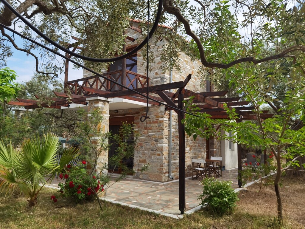 The exterior of the stone house is surrounded by lush greenery, including trees and flowering plants. A natural stone terrace is visible, shaded by a pergola. The balcony on the upper floor features wooden railings, enhancing the traditional architectural style.